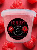 A clear plastic tub filled with pink candy floss, surrounded by fresh red raspberries on a pink background. The front label reads 'RASPBERRY CLOUD 9 CANDY FLOSS' with raspberry and cotton candy graphics.